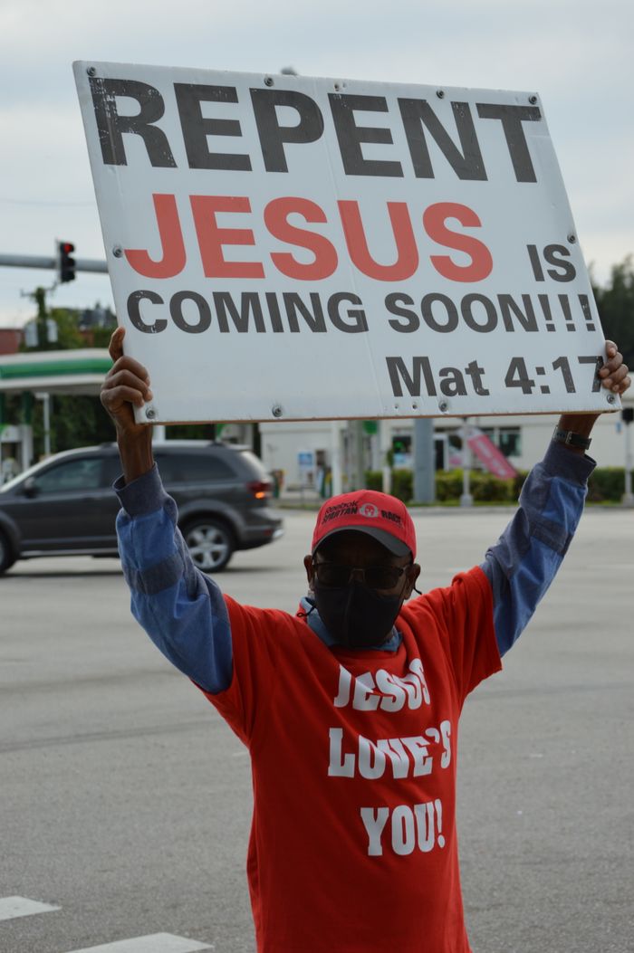 Services Man in red shirt holds a sign with religious message at a busy intersection.